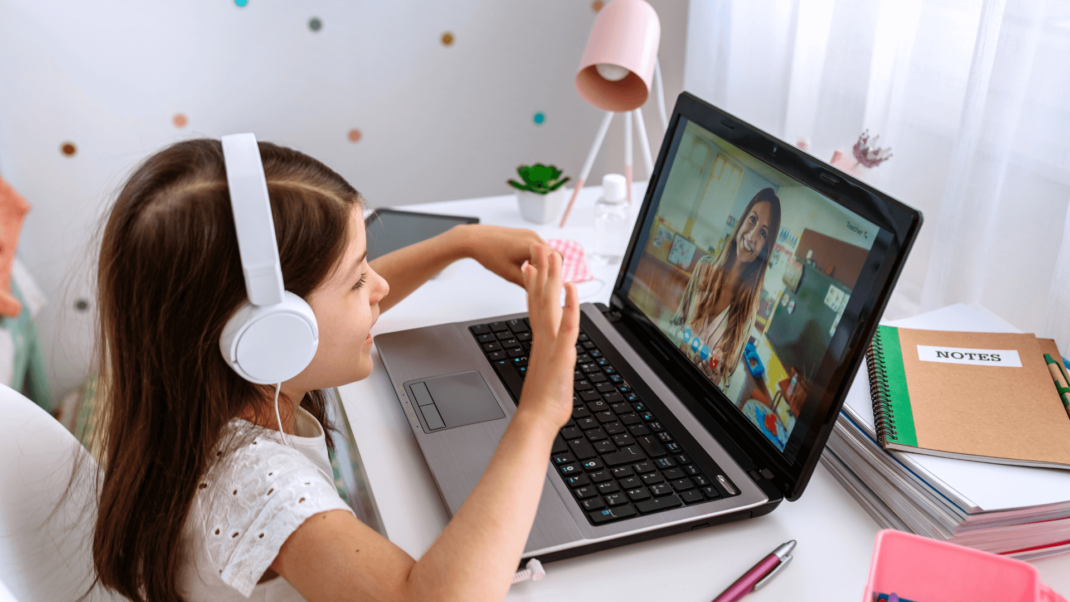 A young girl wearing headphones sits at a desk, waving at a woman on a video call displayed on her laptop.