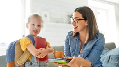 A young girl with a backpack holds a red book while an adult woman hands her notebooks and an apple indoors.