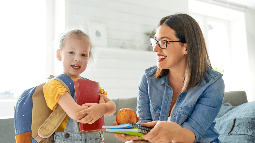 A young girl with a backpack holds a red book while an adult woman hands her notebooks and an apple indoors.
