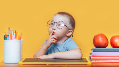A young child wearing glasses sits at a desk with a finger to their lips, surrounded by colored pencils, stacked books, and apples, against a yellow background.