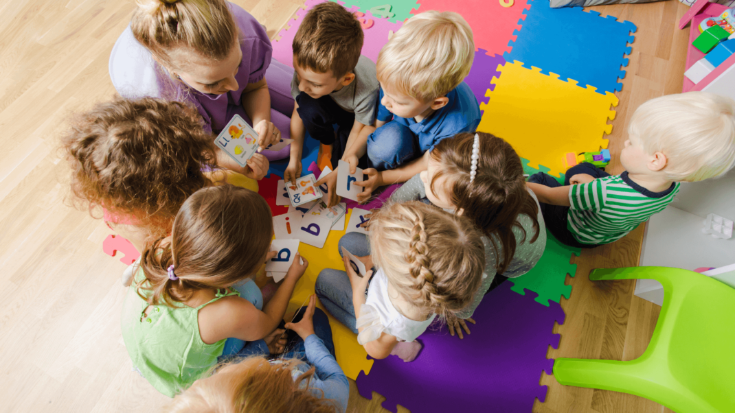 A group of young children sit in a circle on colorful foam mats, looking at alphabet cards with an adult in a classroom or playroom setting.