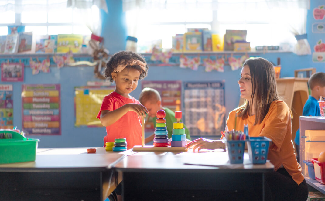 A child stacks colorful rings while a teacher observes at a classroom table, with bright educational materials visible in the background.