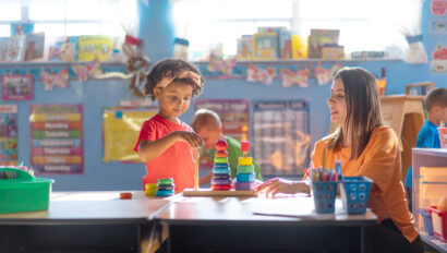 A child stacks colorful rings while a teacher observes at a classroom table, with bright educational materials visible in the background.