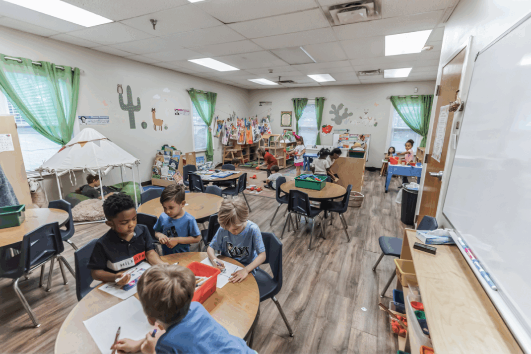 Children sit and work at tables in a classroom decorated with plants and wall art, while others play or read in different activity areas around the room.